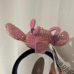 May include: A headband featuring a pink character figure covered in iridescent crystals. The figure has two long, soft pink ears with purple tips and a small gold loop on top. A black headband base supports the figure, and a Disney brand tag is attached.
