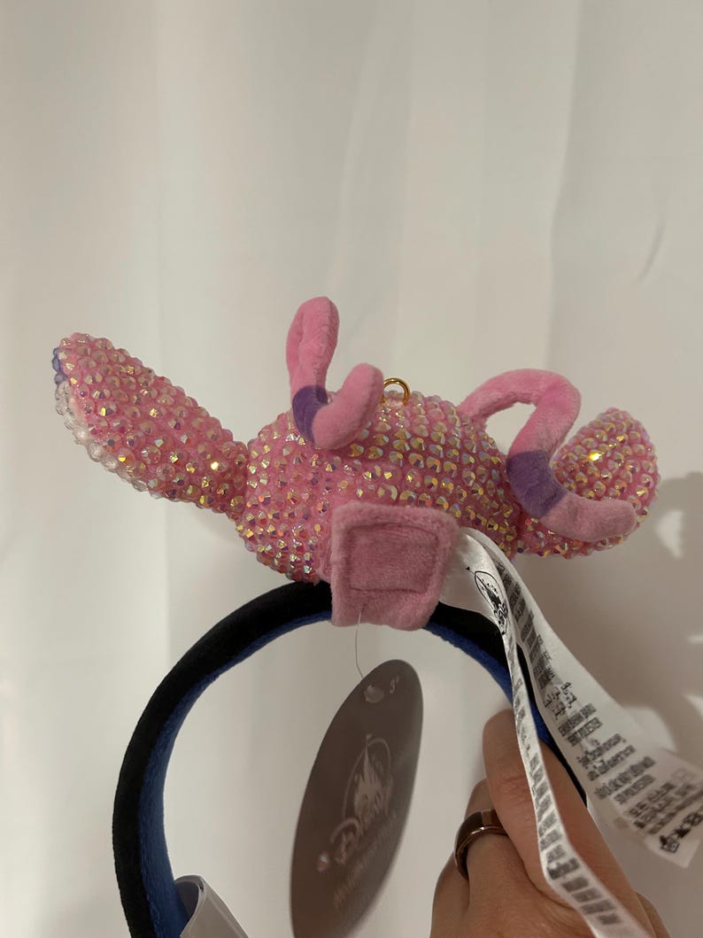 May include: A headband featuring a pink character figure covered in iridescent crystals. The figure has two long, soft pink ears with purple tips and a small gold loop on top. A black headband base supports the figure, and a Disney brand tag is attached.