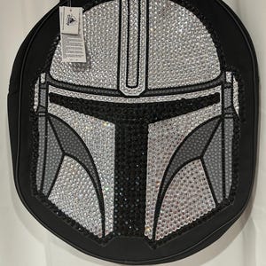 Op de afbeelding: Een ronde zwarte tas met een opvallend Mandalorian-helmdesign, versierd met heldere, grijze en zwarte strass-steentjes. De tas is voorzien van een zwarte schouderband en een wit Disney-merklabel dat met een plastic lusje is bevestigd.