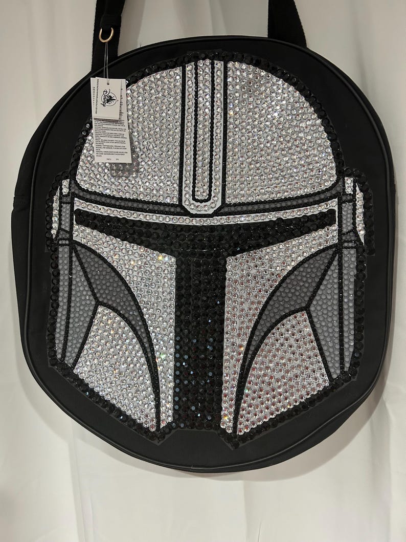 Op de afbeelding: Een ronde zwarte tas met een opvallend Mandalorian-helmdesign, versierd met heldere, grijze en zwarte strass-steentjes. De tas is voorzien van een zwarte schouderband en een wit Disney-merklabel dat met een plastic lusje is bevestigd.
