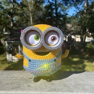 Puede incluir: Una figura amarilla de Minion, adornada con brillantes pedrer&iacute;a, presenta ojos grandes con iris verdes y marrones. La figura lleva un peto azul y guantes negros, con los brazos levantados en un saludo. El fondo muestra una escena soleada al aire libre.