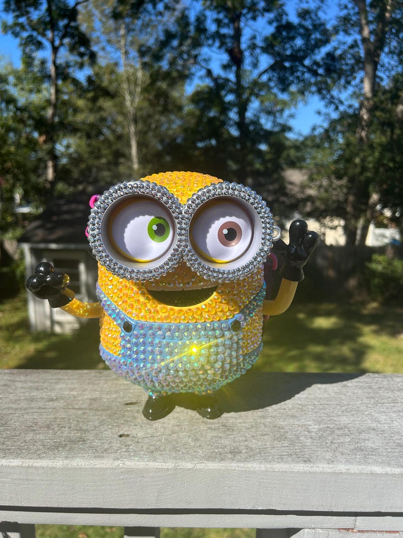 Puede incluir: Una figura amarilla de Minion, adornada con brillantes pedrer&iacute;a, presenta ojos grandes con iris verdes y marrones. La figura lleva un peto azul y guantes negros, con los brazos levantados en un saludo. El fondo muestra una escena soleada al aire libre.
