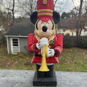May include: A decorative figurine of a marching band leader, featuring a red uniform with gold accents and a black base. The figurine has a Mickey Mouse face and holds a gold trumpet. The figure is covered in sparkling rhinestones.