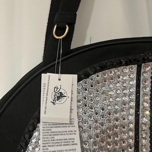 Op de afbeelding: Een zwarte tas met een rond paneel versierd met heldere, reflecterende strass-steentjes en een rand van zwarte pailletten. Een zwarte stoffen riem is bevestigd met een goudkleurige metalen ring. Een wit Disney-label hangt aan de ring met tekst.