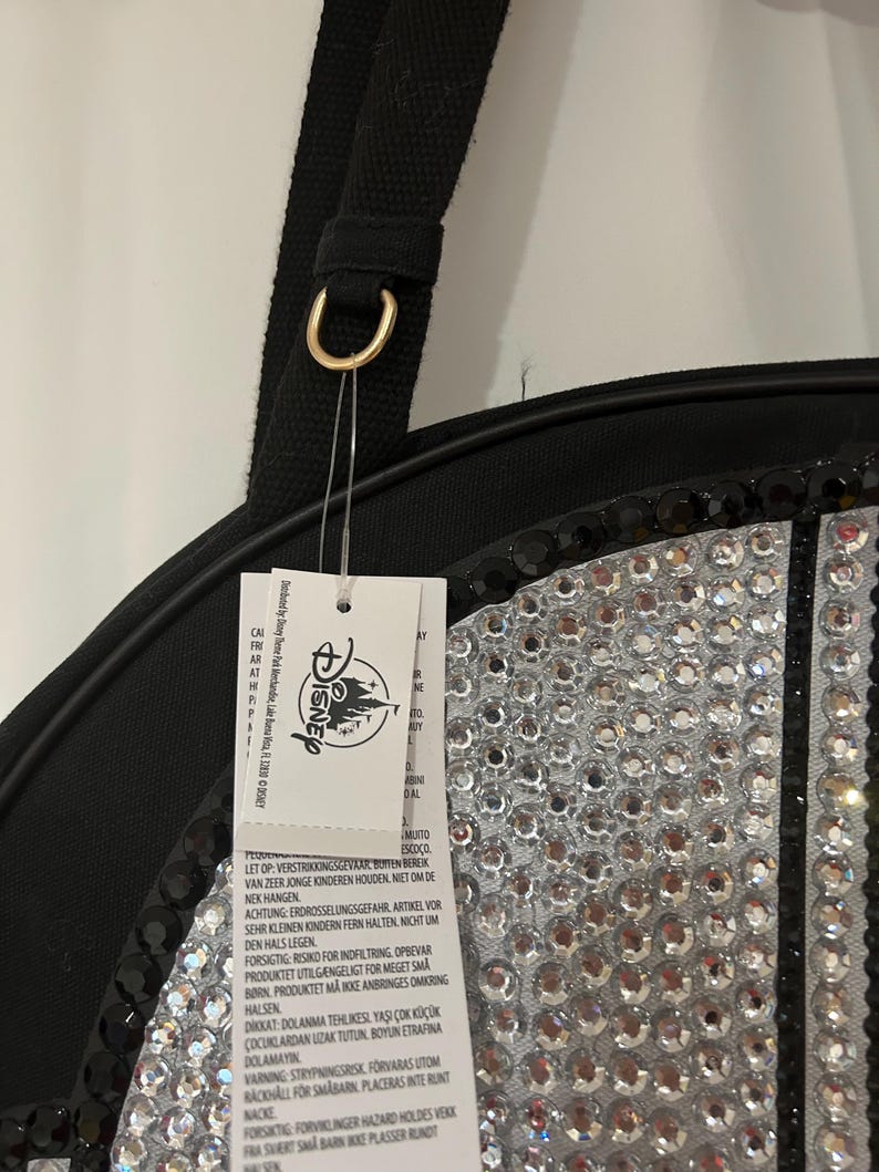 Op de afbeelding: Een zwarte tas met een rond paneel versierd met heldere, reflecterende strass-steentjes en een rand van zwarte pailletten. Een zwarte stoffen riem is bevestigd met een goudkleurige metalen ring. Een wit Disney-label hangt aan de ring met tekst.