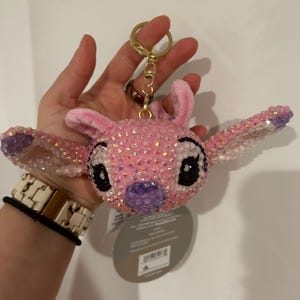 May include: A plush keyring featuring the head of the character Stitch, covered in iridescent pink and purple rhinestones. The character has large ears, black eyes, and a purple nose. A gold-toned metal ring and clasp are attached to the top.