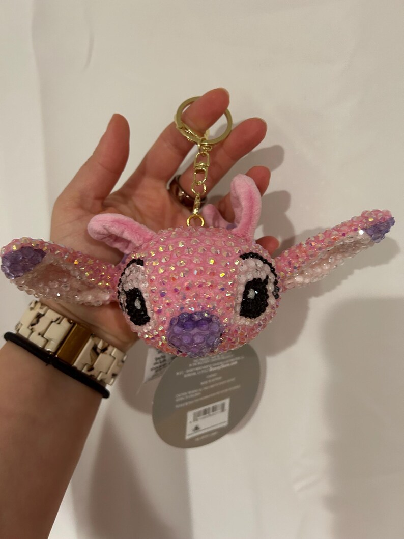 May include: A plush keyring featuring the head of the character Stitch, covered in iridescent pink and purple rhinestones. The character has large ears, black eyes, and a purple nose. A gold-toned metal ring and clasp are attached to the top.