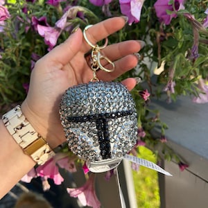May include: A silver and black rhinestone keychain shaped like a helmet, with a gold-colored clasp. The keychain is held in front of a background of purple flowers.