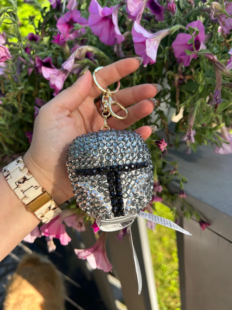 May include: A silver and black rhinestone keychain shaped like a helmet, with a gold-colored clasp. The keychain is held in front of a background of purple flowers.