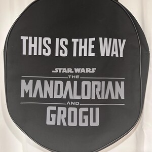 Op de afbeelding: Een zwarte ronde tas met de witte tekst THIS IS THE WAY, STAR WARS THE MANDALORIAN AND GROGU op de voorzijde. De tas is voorzien van zwarte stoffen handvatten aan de bovenkant en een zwarte bies langs de gehele ronde omtrek.