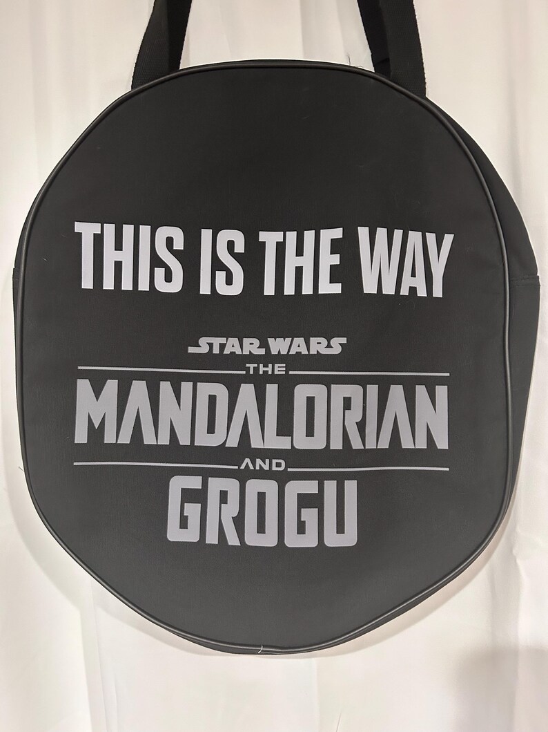 Op de afbeelding: Een zwarte ronde tas met de witte tekst THIS IS THE WAY, STAR WARS THE MANDALORIAN AND GROGU op de voorzijde. De tas is voorzien van zwarte stoffen handvatten aan de bovenkant en een zwarte bies langs de gehele ronde omtrek.