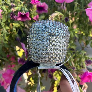 May include: A silver-colored, bedazzled decorative headpiece with a rounded top, adorned with numerous clear rhinestones. The headpiece is attached to a black headband with a blue interior. Pink flowers are in the background.