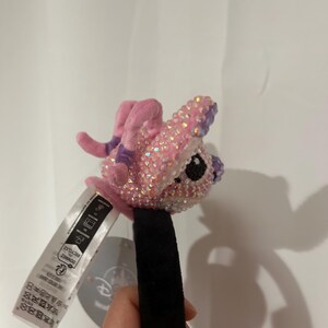 May include: A headband featuring a plush, pink character figure covered in iridescent, light-reflecting crystals. The character has large, dark eyes and small, pink limbs. A white fabric tag with black text and a circular Disney logo tag are attached.