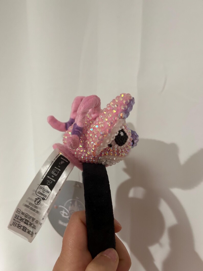 May include: A headband featuring a plush, pink character figure covered in iridescent, light-reflecting crystals. The character has large, dark eyes and small, pink limbs. A white fabric tag with black text and a circular Disney logo tag are attached.