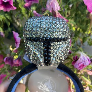May include: A Mandalorian helmet-shaped headband plush, encrusted with sparkling rhinestones. The black and silver design is set against a black and blue headband. A Disney tag is attached.