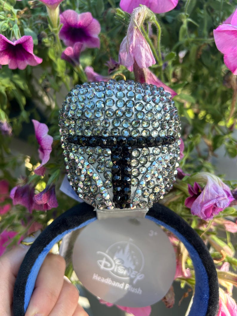 May include: A Mandalorian helmet-shaped headband plush, encrusted with sparkling rhinestones. The black and silver design is set against a black and blue headband. A Disney tag is attached.