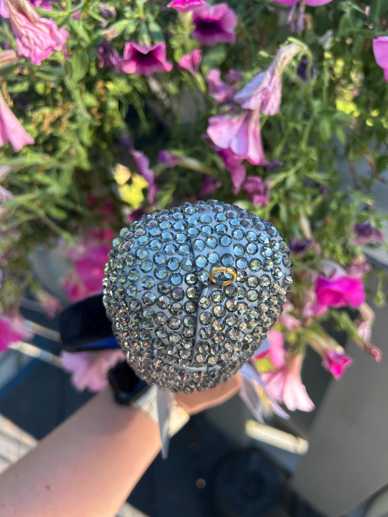 May include: A silver-colored, bedazzled ball-shaped object with a small gold ring attached. The object is held against a backdrop of pink and purple flowers.
