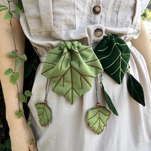 May include: A beige button-down shirt featuring two fabric pouches shaped like leaves attached to the waist. One pouch is light green with brown vein embroidery, and the other is dark green with light green veins, both with dangling leaf charms.