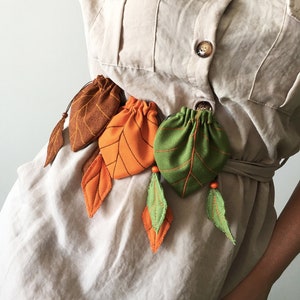 Puede incluir: Un cinturón decorativo con bolsos en forma de hojas en marrón, naranja y verde. Los bolsos están unidos a detalles en forma de hojas con costuras naranjas y verdes. El cinturón está diseñado para usarse alrededor de la cintura.