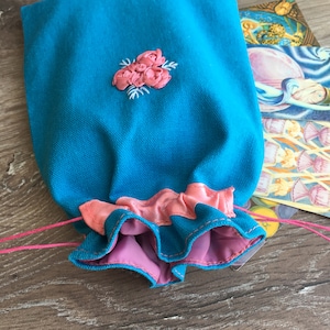 Puede incluir: Una bolsa turquesa con cordón y bordado floral rosa y un borde con volantes. La bolsa tiene un interior rosa y está atada con una cuerda rosa. El fondo incluye una superficie de madera y una tarjeta colorida.