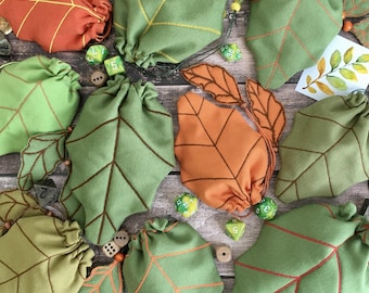 May include: A collection of fabric pouches shaped like autumn leaves in shades of green and orange, featuring stitched vein details and drawstring closures. The pouches are scattered with polyhedral dice and small watercolor leaf illustrations.