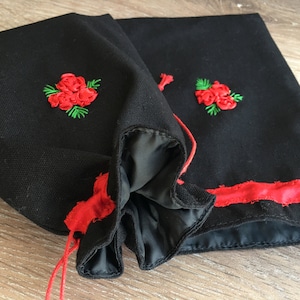 Puede incluir: Dos bolsas de tela negra con ribete de cinta roja y rosas rojas bordadas con hojas verdes. Las bolsas tienen un cierre de cordón con hilo rojo. El interior de las bolsas es negro.