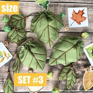 May include: Green leaf-shaped drawstring bags with brown stitching, accompanied by leaf-shaped embellishments and dice. The bags are labeled "SET #3" and "S SIZE". Watercolor illustrations of leaves and an acorn are also present.