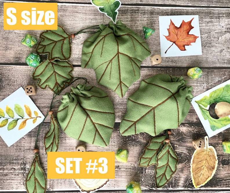 May include: Green leaf-shaped drawstring bags with brown stitching, accompanied by leaf-shaped embellishments and dice. The bags are labeled "SET #3" and "S SIZE". Watercolor illustrations of leaves and an acorn are also present.