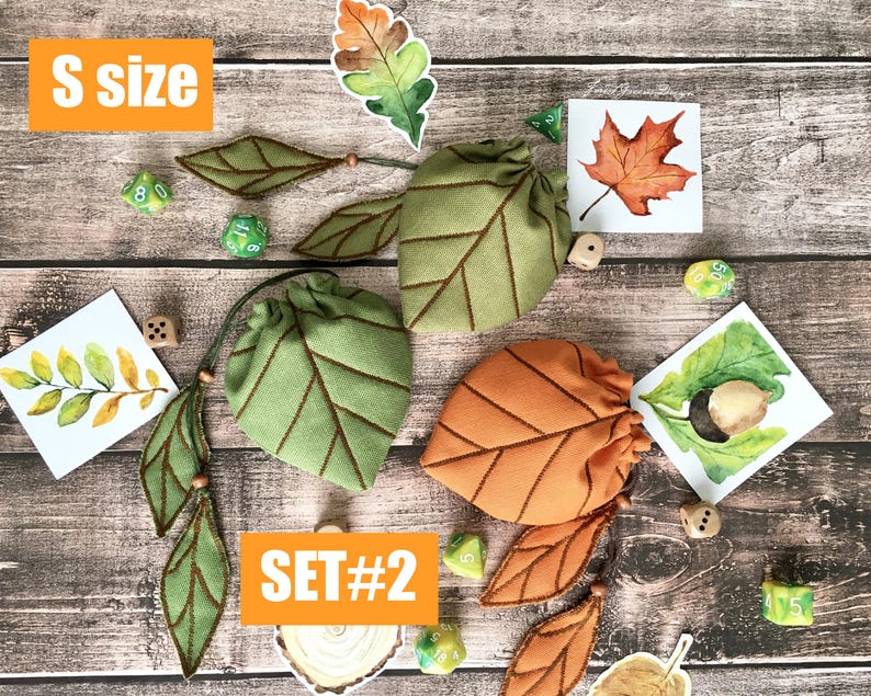 May include: Two leaf-shaped drawstring bags, one green and one orange, with brown stitching, are displayed with leaf-themed embellishments and dice. The text "S size" and "SET#2" are visible. The items are arranged on a wooden surface.