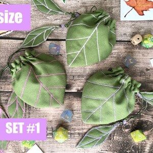 May include: Green leaf-shaped drawstring bags with white and pink embroidered details. The bags are labeled "S size" and "SET #1". Several dice are scattered around the bags, along with leaf-shaped embellishments.