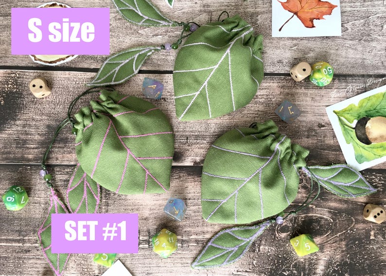 May include: Green leaf-shaped drawstring bags with white and pink embroidered details. The bags are labeled "S size" and "SET #1". Several dice are scattered around the bags, along with leaf-shaped embellishments.