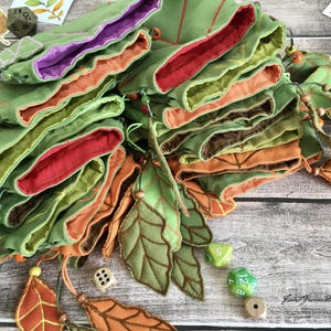 May include: A collection of green fabric dice bags with leaf-shaped appliqu&eacute;s and colorful linings in red, orange, and purple. The bags are arranged in a layered stack, with decorative leaves and dice scattered around.