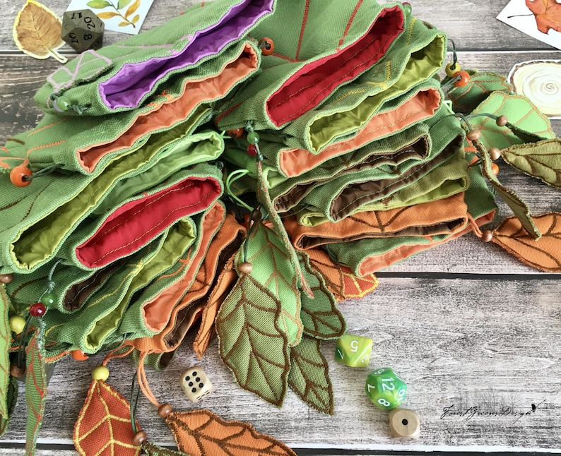 May include: A collection of green fabric dice bags with leaf-shaped appliqu&eacute;s and colorful linings in red, orange, and purple. The bags are arranged in a layered stack, with decorative leaves and dice scattered around.