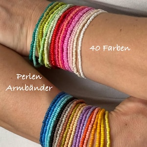 Op de afbeelding: Een stapel kleurrijke kralenarmbanden in verschillende tinten blauw, groen, rood, oranje, geel en roze. De armbanden zijn gestapeld op de pols van een persoon. De tekst "Perlen Armbänder" en "40 Farben" is zichtbaar in de afbeelding.