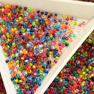 Könnte beinhalten: Eine Sammlung kleiner, durchscheinender, mehrfarbiger Glasperlen in einer Vielzahl lebendiger Farbtöne wie Rot, Blau, Grün, Gelb und Pink. Die Perlen sind auf einer weißen, dreieckigen Sortierschale und einer Oberfläche verstreut.