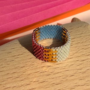 Puede incluir: Un anillo de banda ancha elaborado con pequeñas cuentas en un patrón tejido. El anillo presenta bloques de colores distintos en rojo oscuro, oro metálico, azul claro y blanco hueso. Las cuentas están dispuestas en una cuadrícula estructurada.