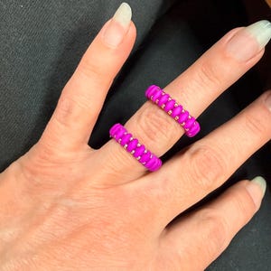 Puede incluir: Dos anillos magenta vibrantes con un diseño trenzado texturizado. Los anillos se usan en los dedos, mostrando su color brillante y textura única. Los anillos son una audaz declaración de moda.