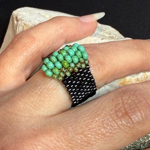 Puede incluir: Anillo de cuentas hecho a mano con una parte superior de cuentas turquesa y verde y una banda de cuentas negras. El anillo se muestra en un dedo, destacando el intrincado trabajo de cuentas y el contraste de color. Una joya única.