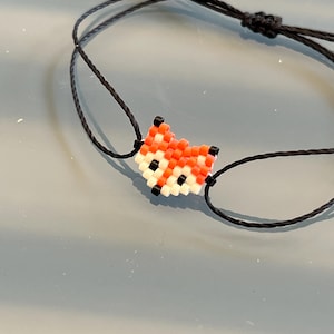 Op de afbeelding: Een verstelbare armband van zwart koord met een rode, witte en zwarte kralenvosbedel.