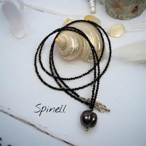 Peut inclure: Un collier en spinelle noir avec un pendentif en perle gris foncé. Le collier est composé de plusieurs rangées de petites perles à facettes. Le mot "Spinell" est écrit en cursive en bas à gauche.