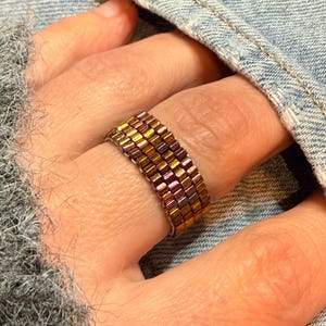 Puede incluir: Un anillo de cuentas con un diseño de mosaico. El anillo presenta pequeñas cuentas rectangulares en tonos dorados, bronce y rosa. El anillo se lleva en un dedo, con una chaqueta vaquera en el fondo.