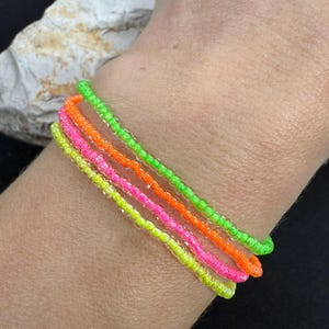 Könnte beinhalten: Vier neonfarbene Armbänder, die auf einem Handgelenk übereinander liegen. Die Armbänder sind grün, orange, pink und hellgrün.