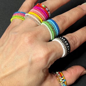 Op de afbeelding: Een hand draagt ​​een stapel kleurrijke kralenringen. De ringen zijn gemaakt van kleine, ronde kralen in verschillende kleuren, waaronder rood, oranje, geel, groen, blauw en paars. De ringen zijn op elkaar gestapeld, waardoor een kleurrijke en opvallende look ontstaat.