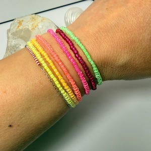 Op de afbeelding: Een verzameling kleurrijke kralenarmbanden in tinten geel, oranje, roze, rood en groen. De armbanden worden samen om een pols gedragen en tonen de levendige kleuren en het delicate kralenwerk. De armbanden zijn gemaakt van kleine, ronde kralen.
