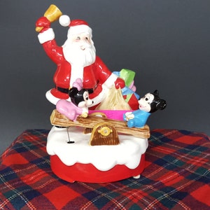 May include: A ceramic Christmas musical figurine featuring Santa Claus ringing a bell, Mickey Mouse and Minnie Mouse on a seesaw. The figurine is decorated with red, white, and green colours.