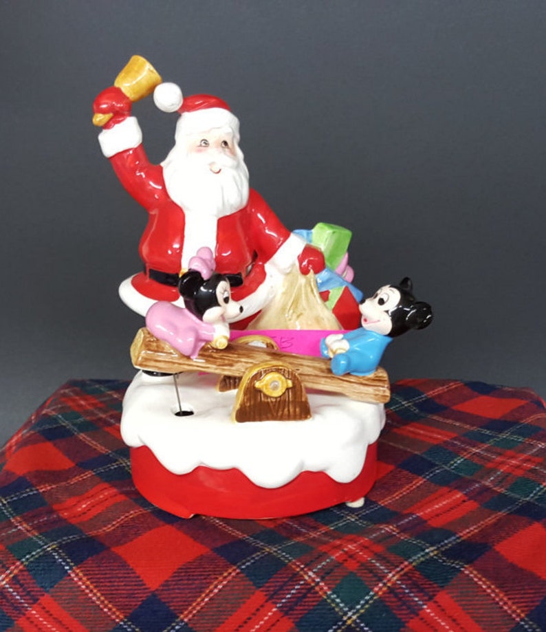 May include: A ceramic Christmas musical figurine featuring Santa Claus ringing a bell, Mickey Mouse and Minnie Mouse on a seesaw. The figurine is decorated with red, white, and green colours.