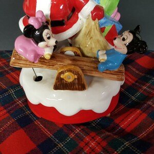 May include: A ceramic Christmas musical figurine of Santa Claus, Mickey Mouse, and Minnie Mouse. Santa is holding a bag of gifts and is on a seesaw with Mickey Mouse. Minnie Mouse is on the other side of the seesaw. The figurine is on a white and red base.