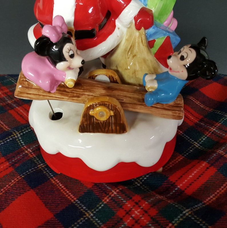 May include: A ceramic Christmas musical figurine of Santa Claus, Mickey Mouse, and Minnie Mouse. Santa is holding a bag of gifts and is on a seesaw with Mickey Mouse. Minnie Mouse is on the other side of the seesaw. The figurine is on a white and red base.