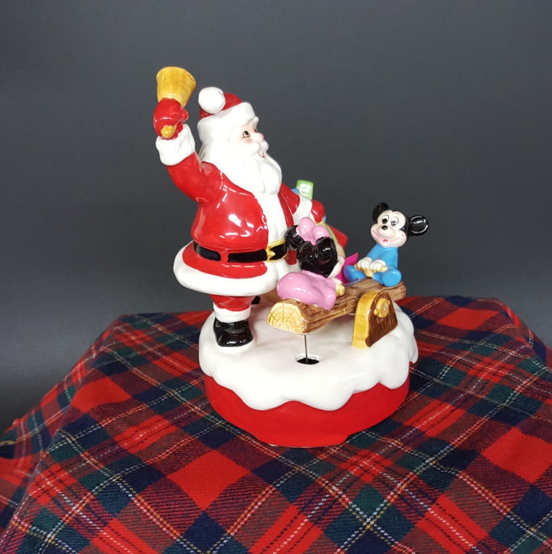 May include: A ceramic Christmas musical figurine featuring Santa Claus ringing a bell, Mickey Mouse and Minnie Mouse on a seesaw. The figurine is on a red and white base with a snowy white top.
