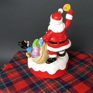 May include: A ceramic Christmas musical figurine of Santa Claus carrying a bag of presents with a small black and blue figure beside him. Santa is wearing a red suit with white trim and a black belt. He is holding a bell in his right hand and is looking to the right. The figurine is on a white base with a red bottom.